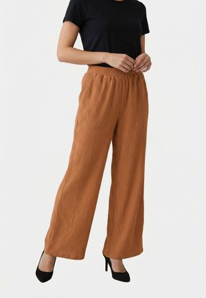 Mujer con pantalones anchos de color óxido, camiseta de manga corta negra y tacones negros, de pie con las manos entrelazadas cerca de la cintura.