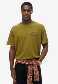 Oliwkowa bawełniana koszulka z krótkim rękawem, z niebieskim nadrukiem logo. W zestawie z ciemnymi dżinsami oraz koszulą w kratkę w odcieniach pomarańczowego i brązowego zawiązaną w talii.