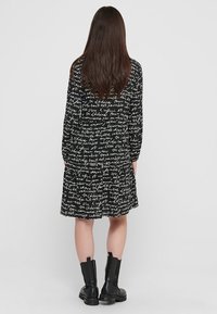 Vestido negro con patrón de texto manuscrito en blanco, mangas largas, corte suelto y dobladillo con volantes, combinado con botas negras hasta el tobillo.