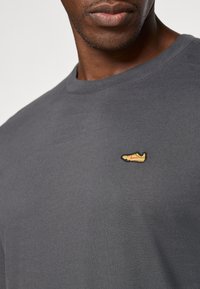 Pilkas medvilnės marškinėliai su apvaliu iškirpimu. Kairėje krūtinės pusėje yra mažas išsiuvinėtas sportbačių logotipas geltonos, raudonos ir juodos spalvų.