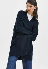 Cardigan oversize di colore blu scuro, realizzato in un morbido maglione a trama. Presenta maniche ampie e un'apertura frontale, abbinato a pantaloni in denim.