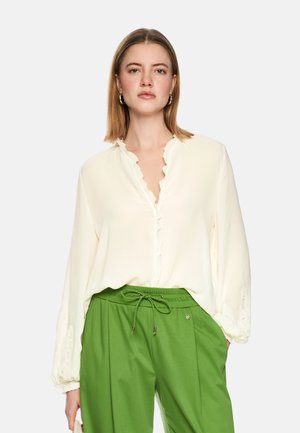 Woman wearing a loose white blouse with ruffled collar and green drawstring pants, standing with hands in pockets against plain background.