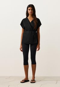 Žena oblečená v čiernom zavinovacom topu s krátkymi rukávmi, čiernych skrátených legínach a čiernych žabkách, stojaca na dlaždicovej podlahe na jednoduchom pozadí.