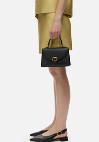 Sac à main en cuir noir avec des finitions dorées, forme structurée, poignée plate et rabat articulé ; associé à un haut doré et des chaussures noires.