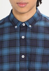 Niebieska i czarna koszula w kratę z kołnierzykiem zapinanym na guziki, z białymi guzikami, kieszenią oraz teksturowanym wzorem materiału.
