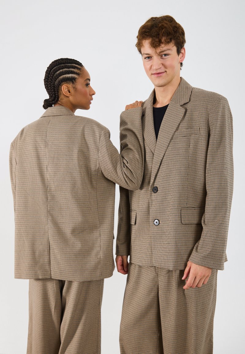 Costume cu carouri maro pe doi modele, caracterizându-se prin sacouri supradimensionate și pantaloni largi. Sacourile au rever cu tăietură și buzunare frontale.