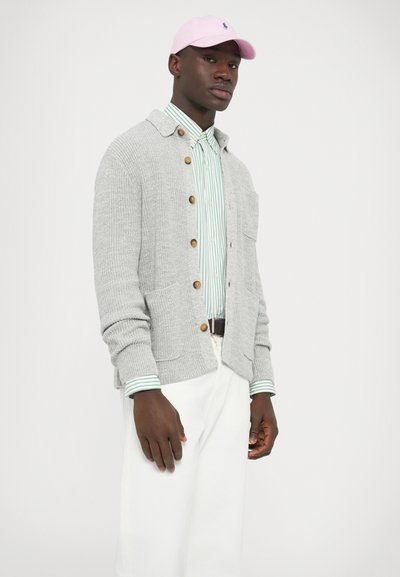 Pilkas megztinis su mediniais mygtukais, dėvimas ant žaliai baltai dryžuoto marškinėlio. Derinamas su baltais kelnais ir rožine kepure.