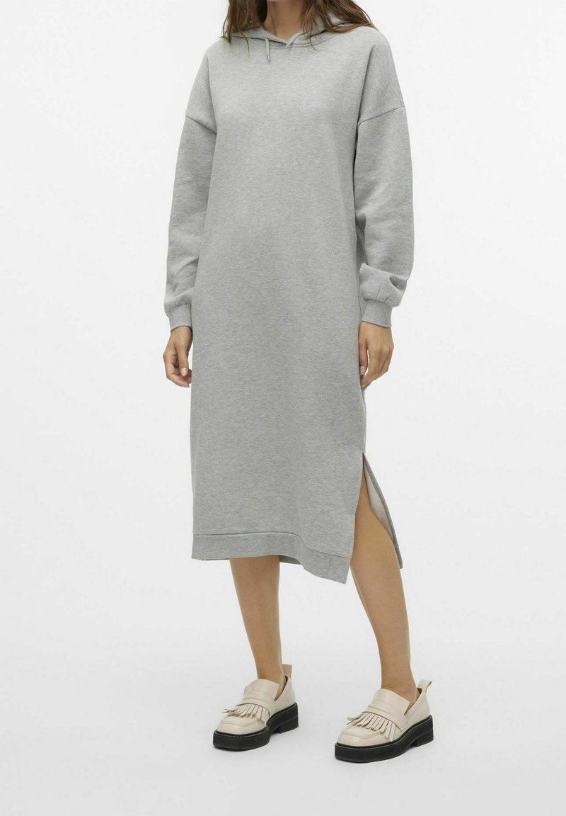 Femme portant une robe midi grise à manches longues avec capuche et fente latérale, associée à des mocassins beiges à semelles noires.