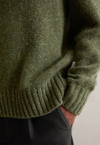 Ελαιογραφί πράσινο πλεκτό πουλόβερ με υφή, με ριγέ μανίκια και στρίφωμα, που διαθέτει διακριτικές πιτσιλιές κίτρινου και λευκού για πρόσθετη λεπτομέρεια.