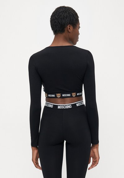 Frau in schwarzem langärmligem Crop-Top und Leggings mit Moschino-Logo-Bändern und Teddy-Bär-Designs, von hinten gezeigt.