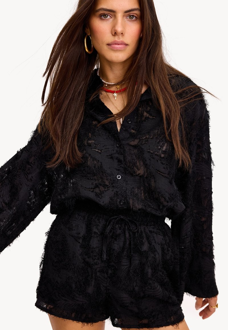 Femme portant un chemisier noir transparent à boutons texturés et un short assorti, accessoirisée avec des boucles d'oreilles créoles en or et des colliers superposés.