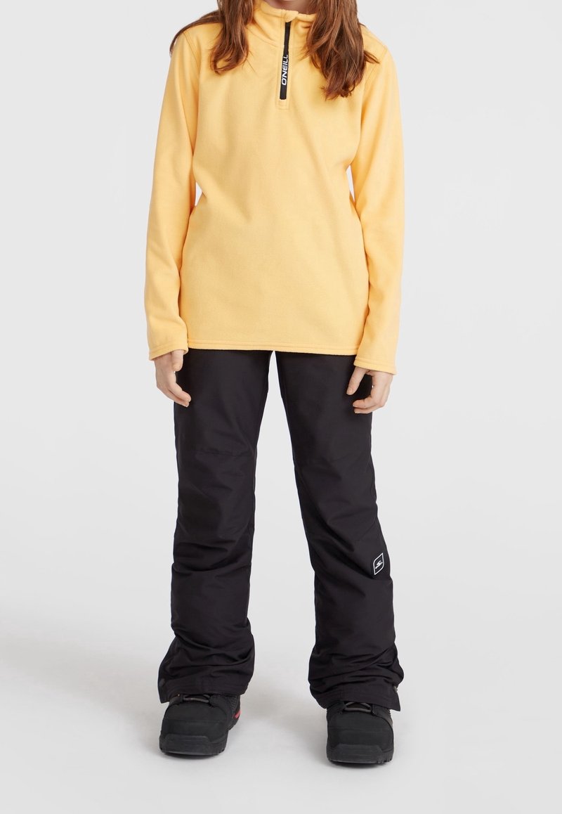 Personne portant un polaire jaune zippé et un pantalon de neige noir, debout sur un fond clair uni.
