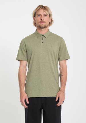 Hombre rubio que lleva una camiseta tipo polo de manga corta color verde oliva claro con botones negros y pantalones negros, de pie sobre un fondo blanco.