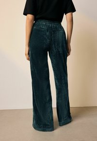 Samtweite-Hosen in dunklem Teal, mit einem elastischen Bund und einer weichenTextur. Auf beiden Seiten sind Gesäßtaschen sichtbar.