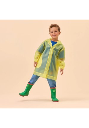 Junges Kind trägt gelben Regenmantel, blaue Jeans und grüne Regenstiefel und steht mit einem Bein angehoben vor einem beigen Hintergrund.