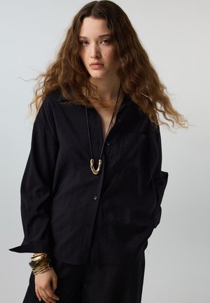 Jeune femme aux longs cheveux bouclés portant une chemise noire ample, un collier en or et plusieurs bracelets en or sur un fond clair uni.