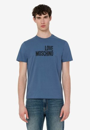 Młody mężczyzna w niebieskim t-shircie z napisem "LOVE MOSCHINO" i niebieskich dżinsach, stojący na jednolitym białym tle.
