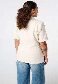 Blouse beige à manches courtes avec une texture lisse et une coupe ajustée, associée à un pantalon large en denim bleu décontracté.