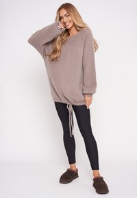 Przesadny, taupe sweter o miękkiej fakturze i ściągaczu na dole, w połączeniu z czarnymi legginsami i brązowymi kapciami, na tle gładkiego białego tła.