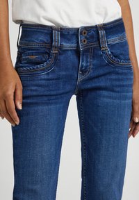 Mitad del cuerpo de una persona que lleva puesta unos jeans de mezclilla azules con cierre de doble botón y costuras naranjas, manos relajadas a los lados.
