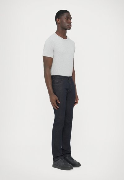 Sivá tričko s krátkym rukávom, tmavomodré úzke rifle a čierne tenisky nosené stojacou osobou, ktorá ukazuje ležérny, moderný outfit.