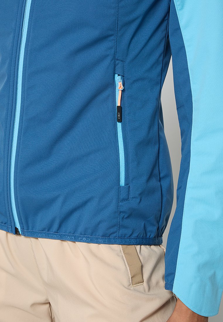 Giacca zip-up blu e azzurro chiaro con tasca laterale, cuciture a contrasto e una texture liscia. Le maniche arrivano al polso.