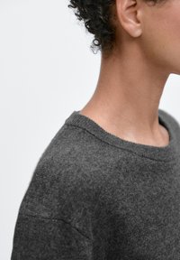 Grauer gestrickter Pullover mit rundem Ausschnitt, weicher Textur, lockerer Passform und sichtbaren Steppnähten nahe Kragen und Schultern.