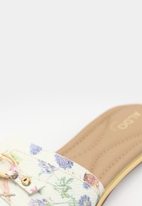 Sandale à enfiler à motif floral avec une tige blanche texturée, une garniture dorée et une semelle intérieure beige rembourrée avec des détails de couture.