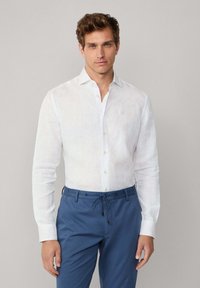 Man in een witte langarmige button-up shirt en blauwe broek met trekkoord, staand tegen een effen lichtgrijze achtergrond.