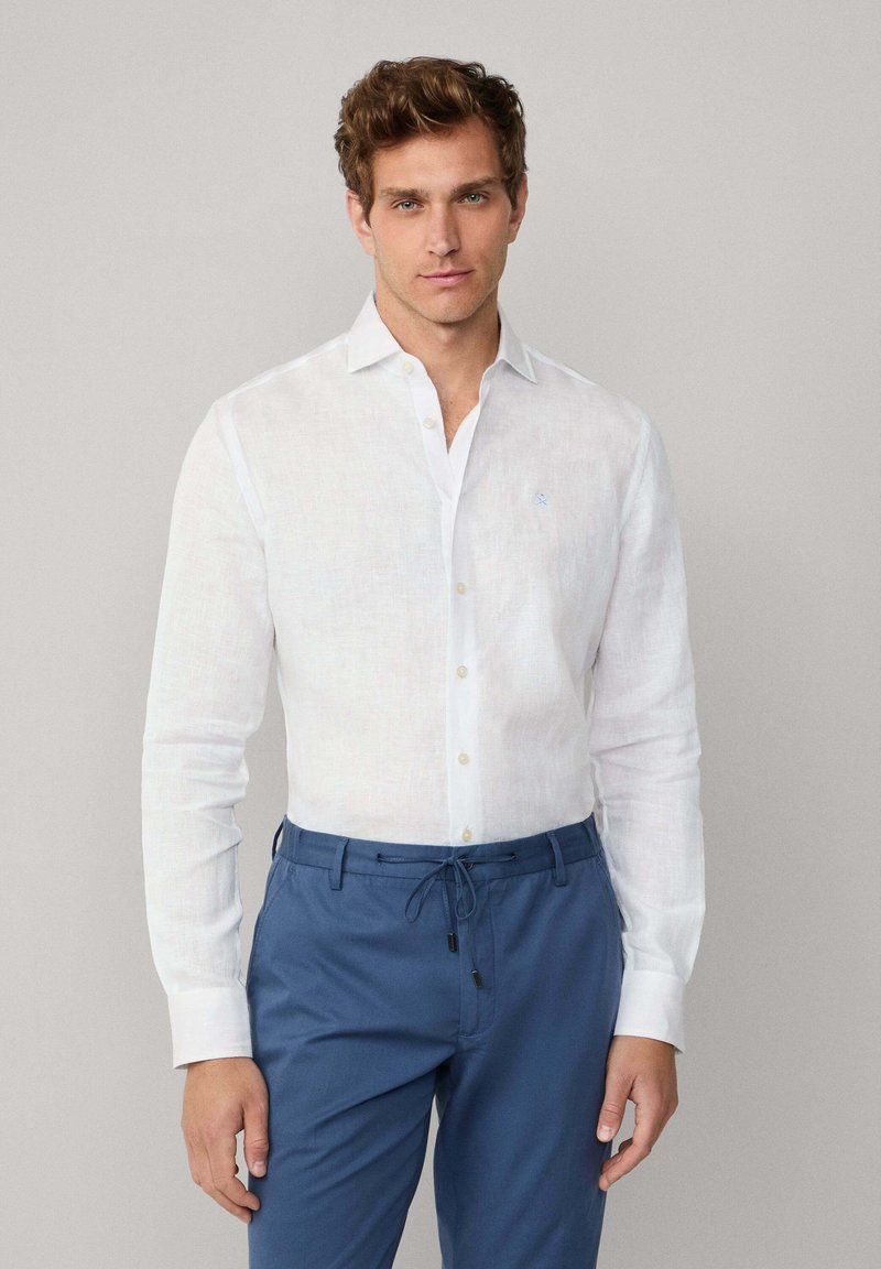 Man in een witte langarmige button-up shirt en blauwe broek met trekkoord, staand tegen een effen lichtgrijze achtergrond.