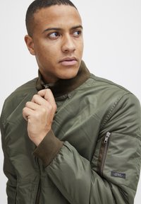 Olive green bomber jacket with a high collar, ribbed cuffs, and a zippered pocket on the sleeve; smooth, shiny fabric texture.