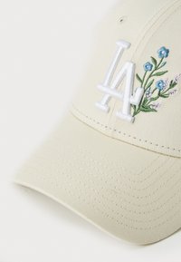 Beige keps med broderad "LA"-logotyp och blommönster. Tillverkad av tyg med en strukturerad form, med en böjd skärm och sydda detaljer.
