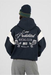 Námornícka mikina s bielymi ramennými panelmi. Na chrbte je text "One Prohibited Social Club" a grafika koňa. Sivé nohavice dopĺňajú outfit.