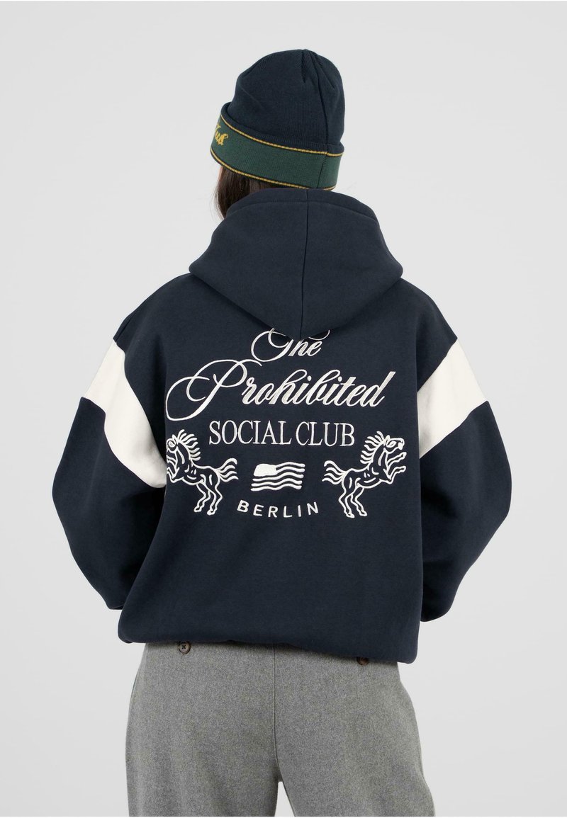 Námornícka mikina s bielymi ramennými panelmi. Na chrbte je text "One Prohibited Social Club" a grafika koňa. Sivé nohavice dopĺňajú outfit.