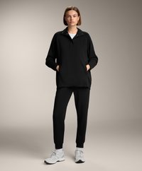 Zwarte capuchonpullover met ritssluiting bij de kraag en zijzakken, gecombineerd met zwarte joggingsbroek. Schoenen: lichtgrijze sportschoenen.