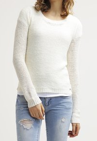 Vit stickad tröja med rundad halsringning, långa ärmar och ribbade mudd. Kombinerad med ljusblå slitna jeans.