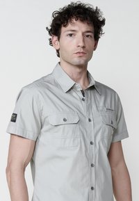 Camicia grigia chiaro a maniche corte con chiusura a bottoni, due tasche sul petto e dettagli neri sui bottoni; presenta una toppa con logo sulla manica.