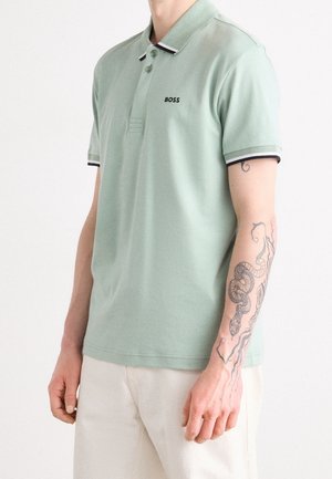 Man wearing light green polo shirt with "BOSS" logo on chest and snake tattoo on left forearm, paired with light-colored pants.