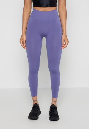 Vrouw draagt paarse leggings met een hoge taille en zwarte sportschoenen, staand tegen een eenvoudige lichte achtergrond.