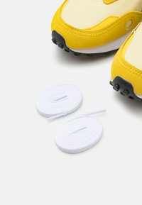 Gelbe Sportschuhe mit einem Obermaterial aus Wildleder und Mesh, ausgestattet mit schwarzen Gummisohlen. Zwei weiße Schnürsenkel befinden sich unter den Schuhen.