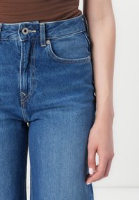 Hoge taille blauwe spijkerbroek met een lichte wassing, zichtbare stiksels, vijf zakken en een knoopsluiting. Hand rustend op de dij.