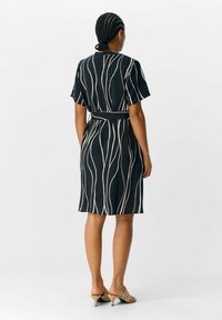 Robe noire avec une taille ajustée, des manches courtes et un tissu texturé. Présente des lignes blanches ondulées. Assortie à des mules à talons beiges.