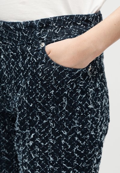 Main partiellement dans la poche d'un pantalon bleu foncé texturé à motif de taches blanches, porté avec un haut blanc.