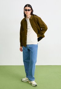 Hnedá kordová košeľa-jacket, biele polokošeľa, modré denimové džínsy a svetlé tenisky s čiernymi slnečnými okuliarami. Zelené pozadie na podlahe.