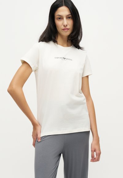 Γυναίκα με σκούρα μαλλιά που φοράει λευκό T-shirt Emporio Armani και γκρι παντελόνι, στέκεται με το ένα χέρι στη μέση της μπροστά από ένα απλό φόντο.