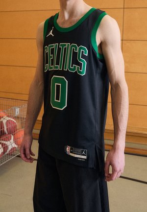 Person trägt ein schwarzes und grünes Boston Celtics Basketballtrikot mit der Nummer Null und steht drinnen neben einem Korb voller Basketbälle.