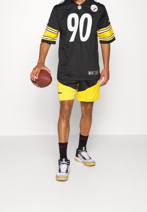 Maillot NFL noir avec des accents jaunes et blancs, numéro 90 ; short jaune et noir ; chaussettes noires avec un logo rouge ; chaussures de sport grises et noires.