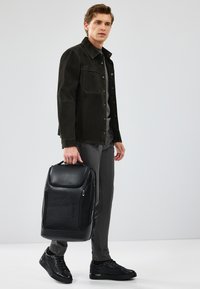 Zaino nero texture con cerniere, portato in una mano. Il modello indossa una giacca scura e pantaloni grigi, in piedi contro uno sfondo neutro.