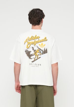 T-shirt branca de algodão com estampa gráfica de um esquiador, cena de montanha e texto "Adidas Originals" em amarelo nas costas.