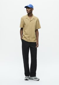 Chemise beige à manches courtes et col, pantalon noir et baskets grises. Comprend une casquette bleue avec un logo. Design épuré, coupe ajustée.